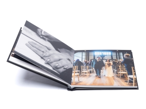 Open photo album displaying wedding photos, left page a close-up of clasped hands, right page guests at a ceremony in chairs.
